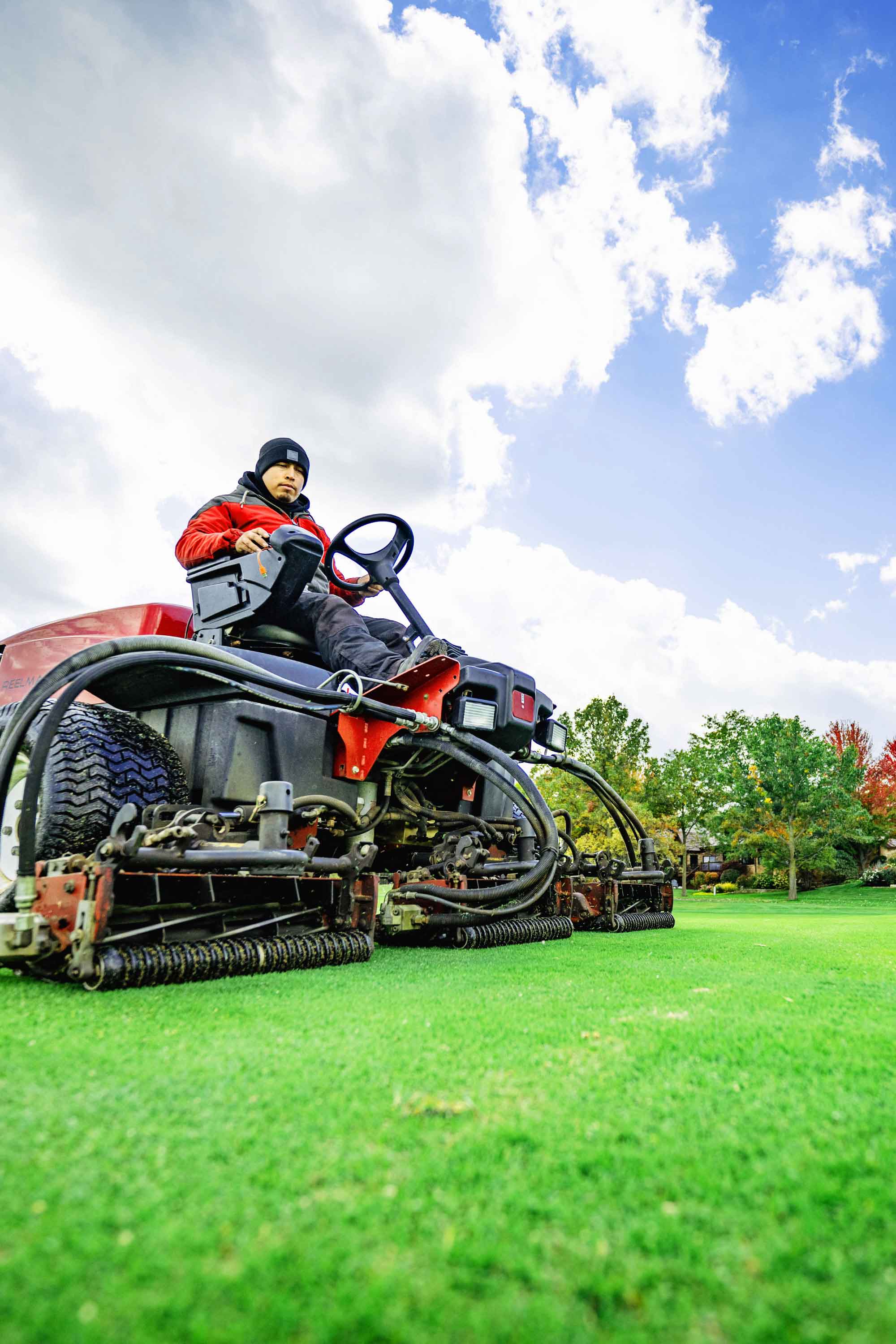 Man on professional mower on fairway cutting grass under a cloudy blue sky