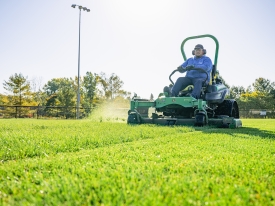 Sports field manager cuts green turf grass on a large riding lawn mower. 