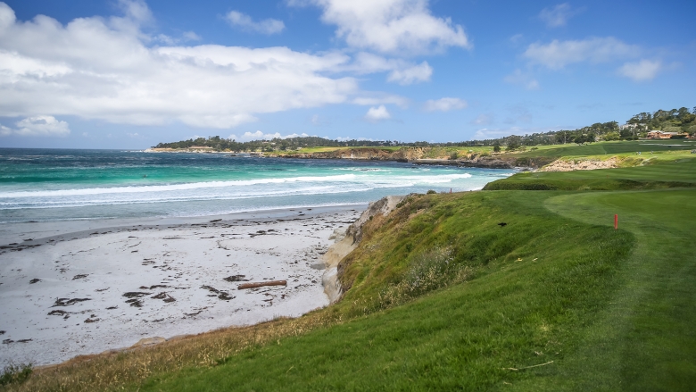 Green golf course rising above a white sandy beach next to blue ocean with white waves 
