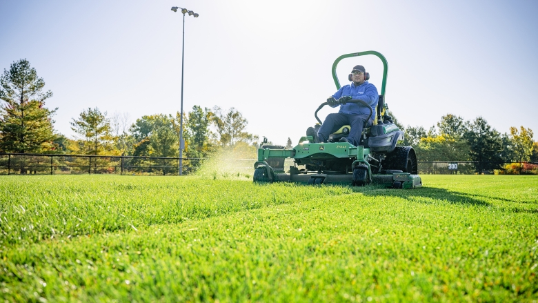 Sports field manager cuts green turf grass on a large riding lawn mower. 