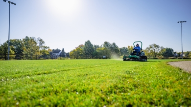 Mower working on the outfield of baseball diamond with trees in the background
