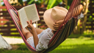 Woman in a hat relaxed in a backyard hammock in the shade while reading a book