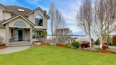 Upscale tan house with green grass trees without leaves in front and a lake in the distance