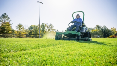 Sports field manager cuts green turf grass on a large riding lawn mower. 