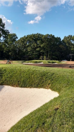 A close up of a golf bunker and sand trap showing green grass with trees in background