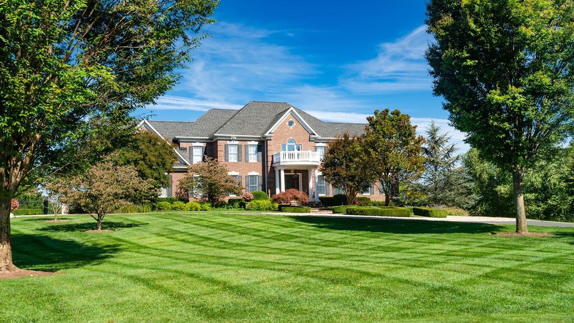 Photo of a high end home with blue sky and green grass in front with trees on each side. 