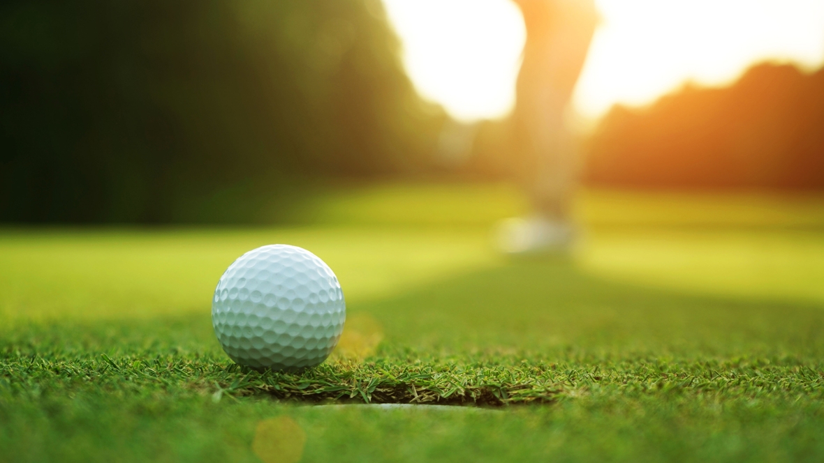 Closeup of a white golf ball next to a putting cup on green grass with blurred golfer in the background