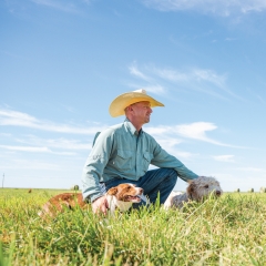 Cowboy with dog sitting in cattle pasture under blue sky.