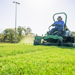 Sports field manager cuts green turf grass on a large riding lawn mower. 